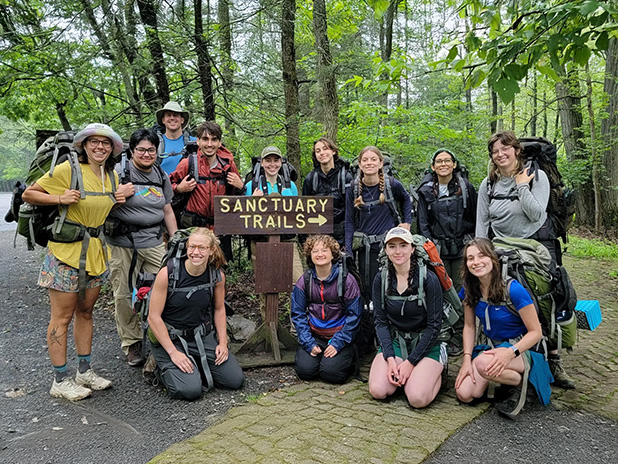 A group of people wearing hiking backpacks pose next to a trail sign and trail.